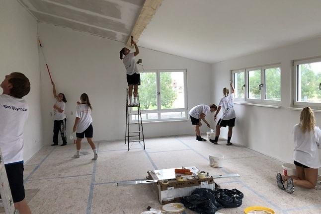 Gruppe von Jugendlichen beim Streichen eines Raums, einige nutzen eine Leiter, andere arbeiten am Boden mit Farbe.