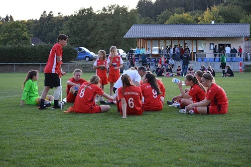 M&auml;dchenteam in roten Trikots sitzt auf dem Rasen, w&auml;hrend ein Trainer ihnen Anweisungen gibt. Zuschauer im Hintergrund.