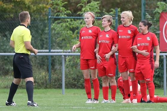 Vier Fu&szlig;ballspielerinnen in roten Trikots stehen in einer Reihe und h&ouml;ren einem Schiedsrichter an einem gr&uuml;nen Fu&szlig;ballfeld zu.