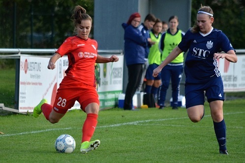 Spielerin in rotem Trikot dribbelt den Ball auf dem Fu&szlig;ballfeld, w&auml;hrend eine Gegnerin sie verfolgt.