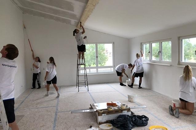 Gruppe von Jugendlichen beim Streichen eines Raums, einige nutzen eine Leiter, andere arbeiten am Boden mit Farbe.