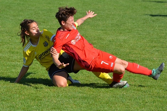 Zwei Fu&szlig;ballspielerinnen k&auml;mpfen am Boden um den Ball auf einem gr&uuml;nen Spielfeld. Eine tr&auml;gt ein rotes, die andere ein gelbes Trikot.