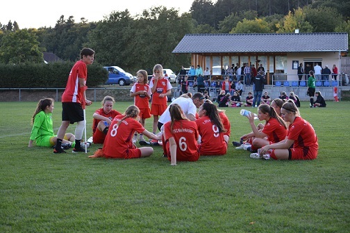 M&auml;dchenteam in roten Trikots sitzt auf dem Rasen, w&auml;hrend ein Trainer ihnen Anweisungen gibt. Zuschauer im Hintergrund.