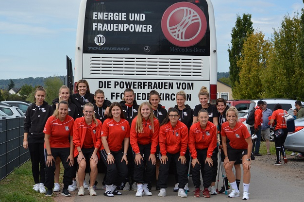 Gruppe von Fu&szlig;ballspielerinnen in roten Trikots posiert vor einem Reisebus mit dem Schriftzug &bdquo;Energie und Frauenpower&ldquo;.