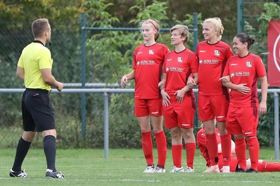 Vier Fu&szlig;ballspielerinnen in roten Trikots stehen in einer Reihe und h&ouml;ren einem Schiedsrichter an einem gr&uuml;nen Fu&szlig;ballfeld zu.