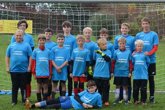 Gruppe von 14 Jungen in blauen T-Shirts mit der Aufschrift &bdquo;Fu&szlig;ballcamp&ldquo; vor einem Fu&szlig;balltor auf dem Rasen.