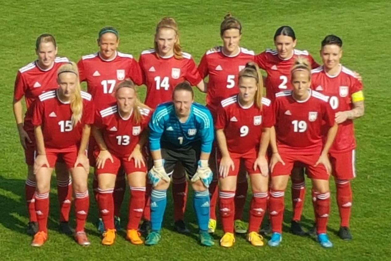 Frauenfußballmannschaft in roten Trikots posiert auf dem Spielfeld, mit einer Torhüterin in blauer Ausrüstung in der Mitte.