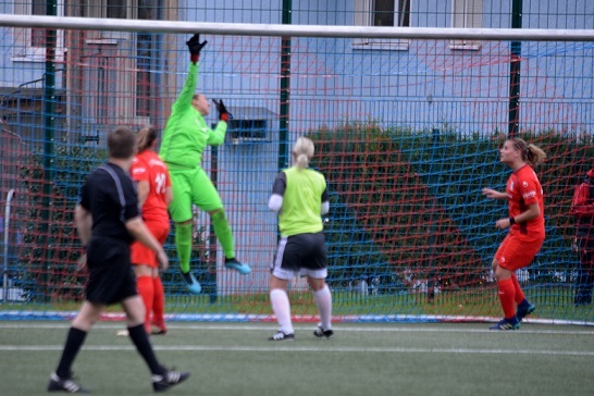Torh&uuml;terin in gr&uuml;ner Ausr&uuml;stung springt, um einen Ball im Fu&szlig;ballspiel zu halten, w&auml;hrend Spielerinnen in roten Trikots agieren.
