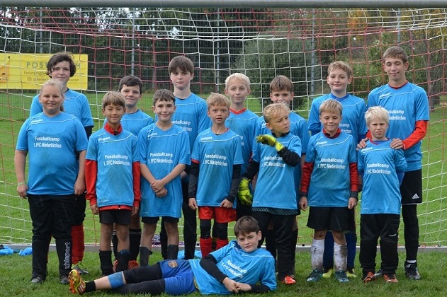 Gruppe von 14 Jungen in blauen T-Shirts mit der Aufschrift &bdquo;Fu&szlig;ballcamp&ldquo; vor einem Fu&szlig;balltor auf dem Rasen.