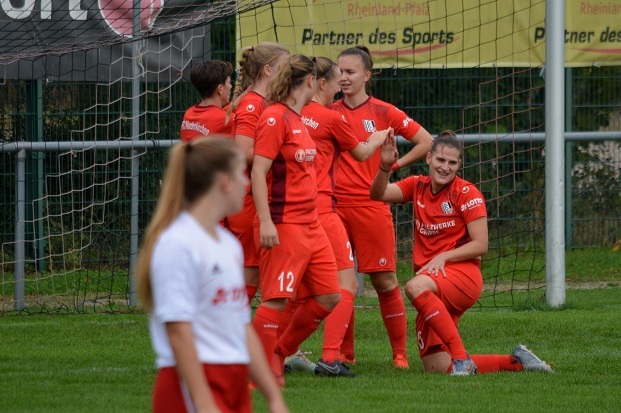 Gruppenkonversation von Fu&szlig;ballspielerinnen in roten Trikots auf einem Sportplatz, eine Spielerin kniet l&auml;chelnd am Boden.