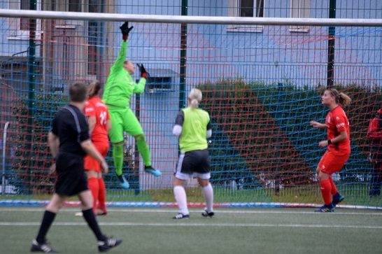 Torh&uuml;terin in gr&uuml;ner Ausr&uuml;stung springt, um einen Ball im Fu&szlig;ballspiel zu halten, w&auml;hrend Spielerinnen in roten Trikots agieren.