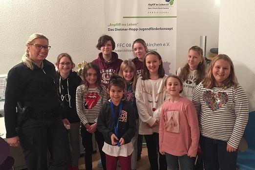 Gruppe von zw&ouml;lf Kindern und einer Erwachsenen vor einem Banner des Dietmar-Hopp-Jugendf&ouml;rderkonzepts in einem Raum.