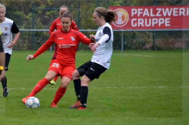 Zwei Fu&szlig;ballspielerinnen im Wettkampf um den Ball auf einem Rasenfeld, im Hintergrund steht ein Werbebanner.