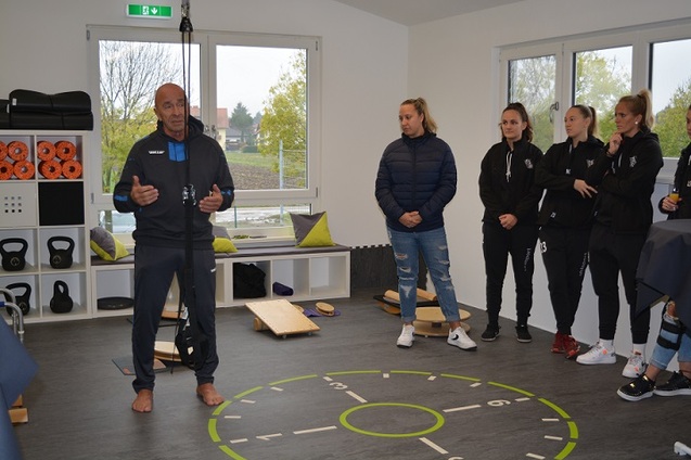 Trainer erkl&auml;rt eine &Uuml;bung in einem modern ausgestatteten Trainingsraum, w&auml;hrend Sportlerinnen aufmerksam zuh&ouml;ren.