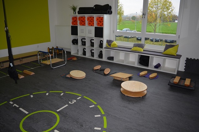 Bewegungsraum mit verschiedenen &Uuml;bungsger&auml;ten, darunter Gleichgewichtsplattformen, Matten und Hanteln auf einem grauen Boden.