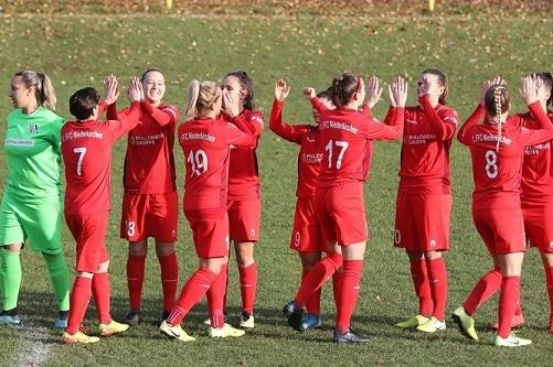 Acht Spielerinnen in roten Trikots klatschen sich auf einem Sportplatz ab, eine Torh&uuml;terin steht in gr&uuml;ner Ausr&uuml;stung daneben.