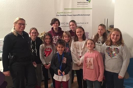 Gruppe von zw&ouml;lf Kindern und einer Erwachsenen vor einem Banner des Dietmar-Hopp-Jugendf&ouml;rderkonzepts in einem Raum.