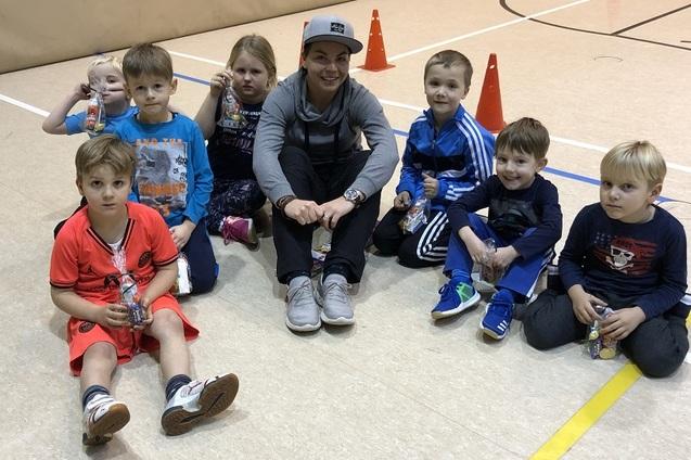 Gruppe von sieben Kindern sitzt auf dem Boden einer Sporthalle, l&auml;chelnd, mit einer Erwachsenen in sportlicher Kleidung.
