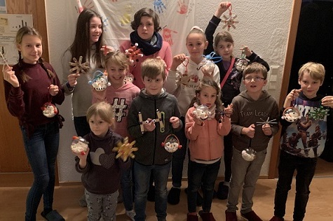 Gruppe von Kindern und Jugendlichen h&auml;lt selbstgemachte Weihnachtsdekorationen vor einer Wand mit Weihnachtsmotiven.