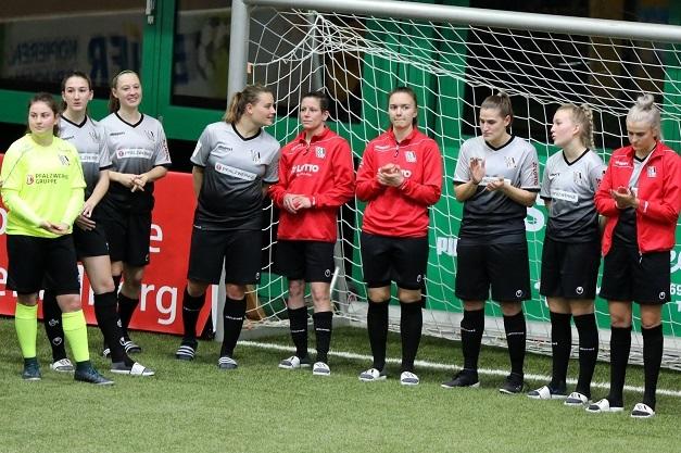 Frauenfu&szlig;ballmannschaft steht in Sportbekleidung am Spielfeldrand, applaudierend vor einem Tor.