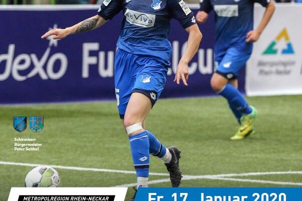Fu&szlig;ballspiel in der Halle mit weiblichen Spielerinnen in blauen Trikots, Publikum im Hintergrund. Veranstaltungshinweis sichtbar.