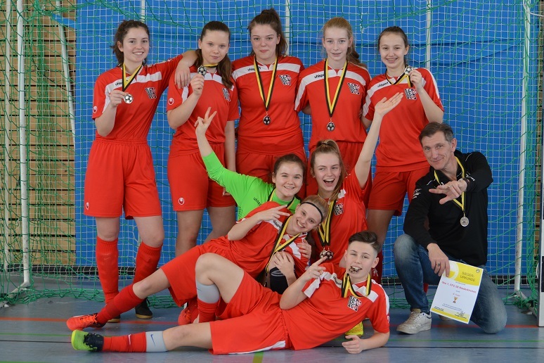 Gruppe von neun Fu&szlig;ballspielerinnen in roten Trikots mit Medaillen, posierend auf einem Sportfeld.