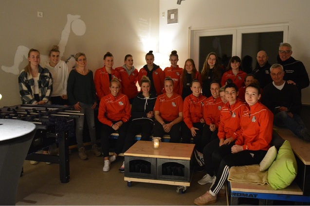 Gruppenfoto von Sportteam und Unterst&uuml;tzern in einem gem&uuml;tlichen Raum, alle in roten Trainingsjacken, mit Tischfu&szlig;ball.
