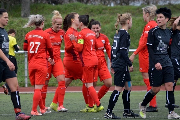Spielszene mit Spielerinnen in roten und schwarzen Trikots, die nach einem Tor am Rasenfeld feiern und sich umarmen.