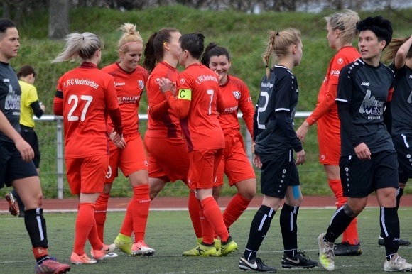 Spielszene mit Spielerinnen in roten und schwarzen Trikots, die nach einem Tor am Rasenfeld feiern und sich umarmen.