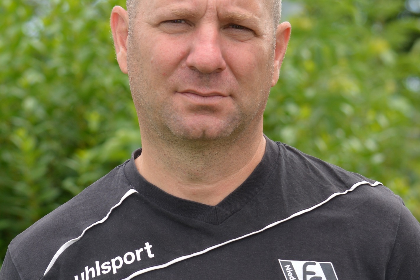 Portr&auml;t eines Mannes in schwarzem Sporttrikot mit Logo, im Freien vor gr&uuml;nen Str&auml;uchern, mit ernstem Gesichtsausdruck.