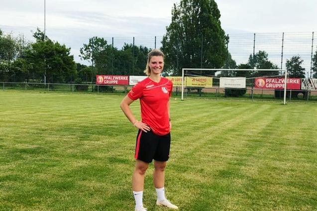 L&auml;chelnde Fu&szlig;ballspielerin in rotem Trikot steht auf einem Rasenplatz neben einem Fu&szlig;balltor.