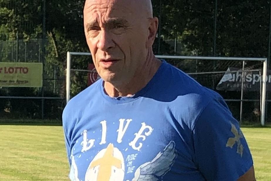 Mann mit Glatze tr&auml;gt blaues T-Shirt und steht auf einem Fu&szlig;ballplatz im Freien. Hintergrund mit Toren und gr&uuml;ner Wiese.