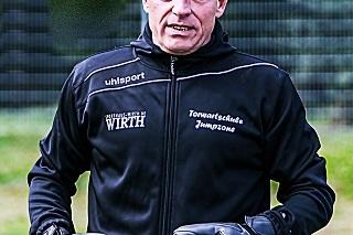 Mann in schwarzer Sportjacke mit Torwarthandschuhen steht auf einem Platz, Hintergrund mit verschwommenem Zaun.