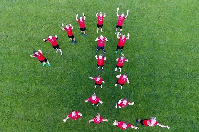 Gruppierung von 17 Personen in roten T-Shirts, die auf gr&uuml;nem Rasen stehen und die Arme in die Luft heben.