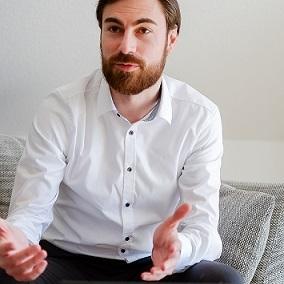 Mann im weißen Hemd sitzt auf einer Couch und spricht mit gestikulierenden Händen, Laptop vor ihm.