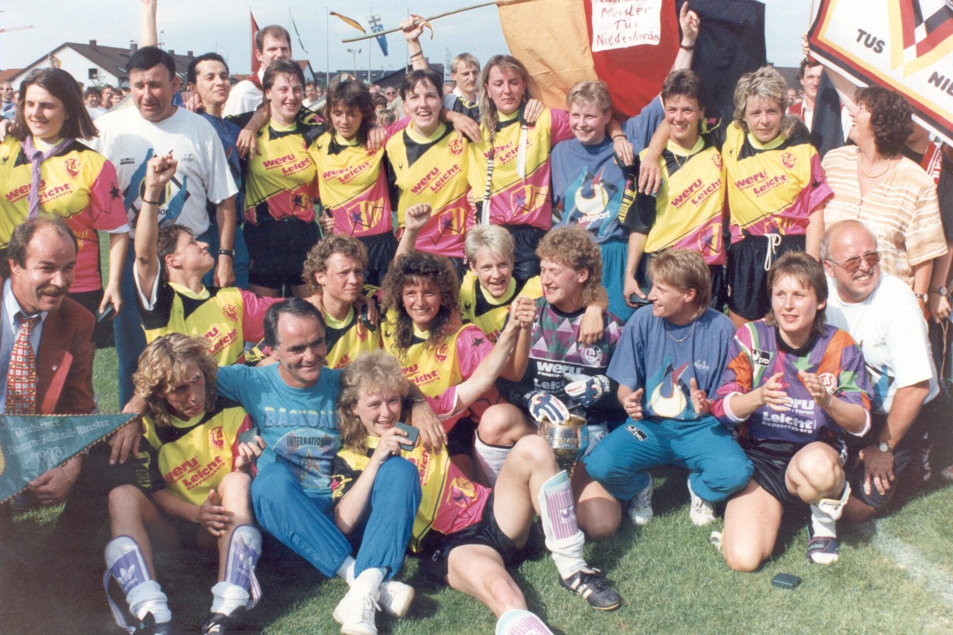 Gruppenshot einer Fu&szlig;ballmannschaft in bunten Trikots, umgeben von Freudenjubel auf einem Sportplatz.