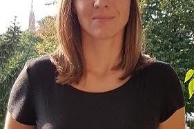 Porträt einer Frau mit schulterlangen Haaren, die in einem schwarzen Shirt vor einer grünen Gartenlandschaft steht.