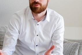 Mann im weißen Hemd sitzt auf einer Couch und spricht mit gestikulierenden Händen, Laptop vor ihm.