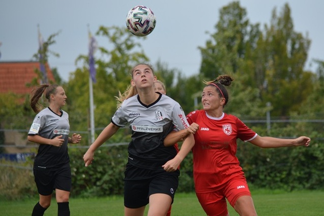 Zwei Fu&szlig;ballspielerinnen im Duell, w&auml;hrend der Ball hoch in die Luft fliegt, im Hintergrund ein Fu&szlig;ballfeld.