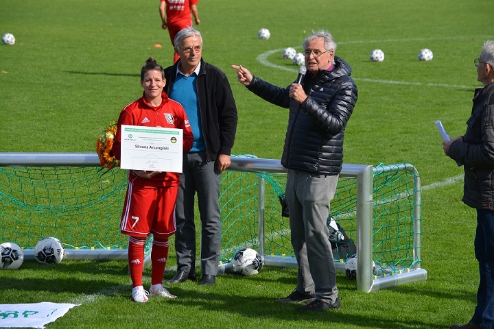 Frau in rotem Fu&szlig;balltrikot mit Auszeichnung in der Hand steht neben zwei M&auml;nnern auf dem Fu&szlig;ballplatz.
