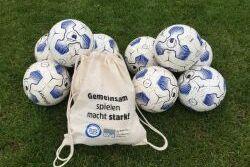 Zehn weiße Fußbälle mit blauen Mustern liegen auf Rasen, neben einem Beutel mit dem Aufdruck „Gemeinsam spielen macht stark!“.