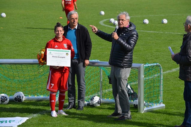 Frau in rotem Fu&szlig;balltrikot mit Auszeichnung in der Hand steht neben zwei M&auml;nnern auf dem Fu&szlig;ballplatz.
