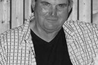 Schwarzwei&szlig;aufnahme eines Mannes mit kurzem, grauem Haar und kariertem Hemd vor einer Holzwand.