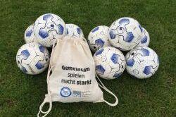 Zehn wei&szlig;e Fu&szlig;b&auml;lle mit blauen Mustern liegen auf Rasen, neben einem Beutel mit dem Aufdruck &bdquo;Gemeinsam spielen macht stark!&ldquo;.