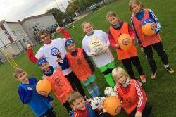 Gruppierung von zehn Kindern in Sportkleidung, die auf einem Rasenplatz bunte B&auml;lle und einen Pokal halten.
