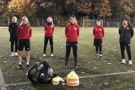 Acht Personen in Sportkleidung stehen auf einem Fu&szlig;ballfeld, umgeben von herbstlichem Laub und Trainingsmaterial.