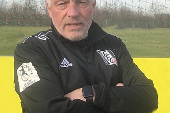 Trainer im Sportanzug mit verschr&auml;nkten Armen vor einem Fu&szlig;balltor und gr&uuml;nem Rasenfeld.