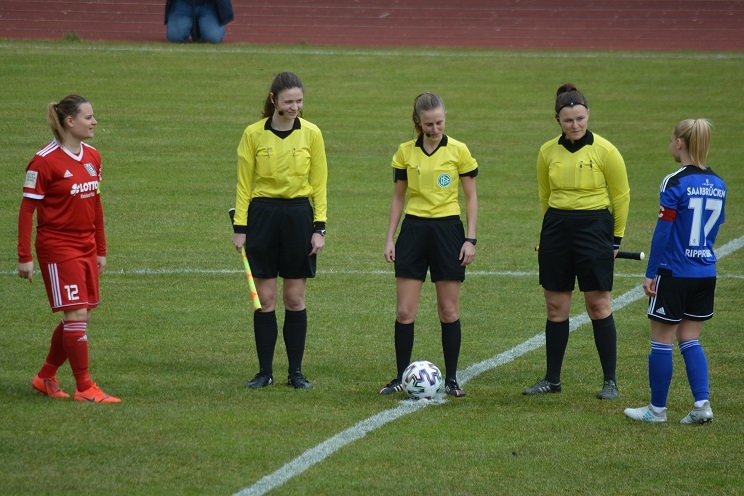 Zwei Fu&szlig;ballspielerinnen und vier Schiedsrichterinnen stehen auf dem Spielfeld zur Spielanpfiff bereit.