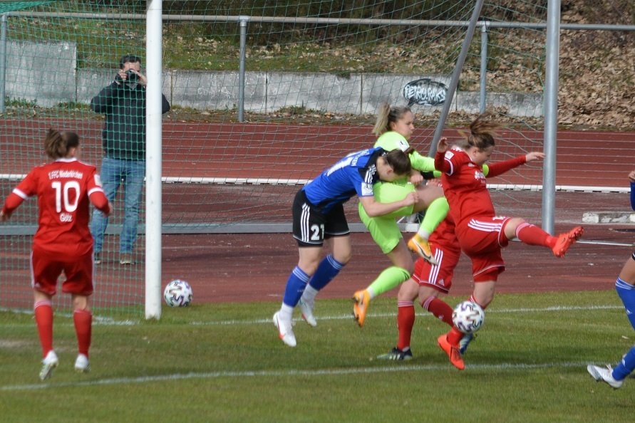 Zwei Fu&szlig;ballspielerinnen k&auml;mpfen im Strafraum um den Ball, w&auml;hrend die Torh&uuml;terin eingreift. Zuschauer im Hintergrund.