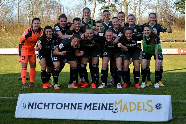 Gruppenfoto einer Frauenfu&szlig;ballmannschaft in schwarzen Trikots, posierend vor einem Banner mit dem Text &bdquo;NICHT OHNE MEINE M&Auml;DELS&ldquo;.